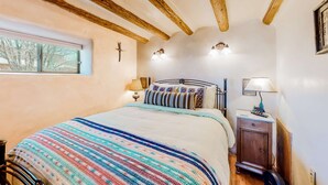 Ein Schlafzimmer mit einem Bett, Holzbalken, einem Fenster, einem Nachttisch mit Lampe und einer Wandleuchte.