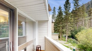 A balcony with a wooden floor, a small stool, and a view of a forested area. - Sierra Del Sol 30 - Hot Tub, Walk to Lifts & Town (Taos Ski Valley)