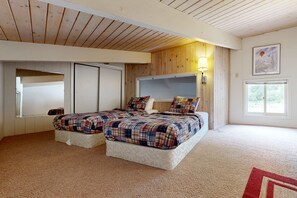 A bedroom with two beds, wooden ceiling, a large window, and a wall-mounted lamp.