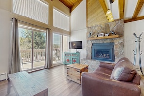 A living room with a stone fireplace, a television, a leather sofa, and a wooden table. - Timbers Five - Walk to Ski, Hike, Bike & Dining (Angel Fire)