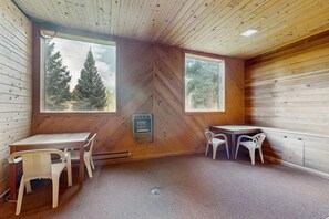 A cabin interior with two windows, a wooden ceiling, a table with chairs, and a small heater. - Ski Peaks Escape - Walk or Shuttle to Ski Lifts (Angel Fire)