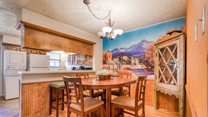 A kitchen with wooden cabinets, a dining table with chairs, and a mural of a mountain landscape. - Creekside Serenade - Hot Tubs & Close to Town (Red River)
