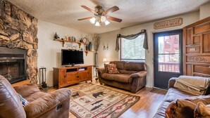 A cozy living room with a stone wall, a fireplace, a television, leather sofas, a patterned rug, and a sign reading 'Friends & Family Gather Here'. - Creekside Serenade - Hot Tubs & Close to Town (Red River)