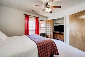 Un dormitorio con cama, ventilador de techo, televisor, puerta de madera y ventana con cortinas rojas.