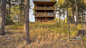 A two-story wooden cabin with a balcony, surrounded by tall pine trees and dry grass. - Bigfoot Lodge - Mountain Views & Multiple Decks (Angel Fire)