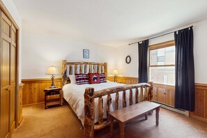 A bedroom with a wooden bed, a bench, a nightstand, a window with curtains, and a wall-mounted picture. - Snowfire 205A - Walk to Ski, Hike, Bike & Dining (Angel Fire)
