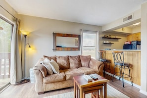A living room with a sofa, a coffee table, a floor lamp, and a kitchen area with bar stools. - Christi Villa 100 - Near Ski, Hike, Bike & Dining (Angel Fire)