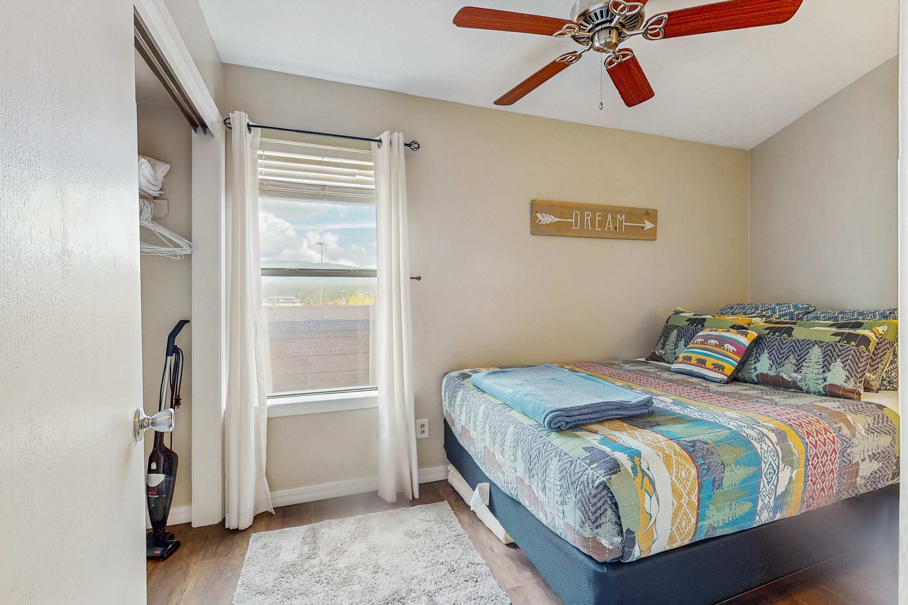 Une chambre à coucher avec un lit, un ventilateur de plafond, une fenêtre avec des rideaux, un aspirateur et un panneau accroché au mur.