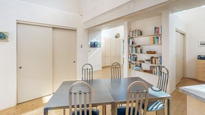 Uma sala de jantar com mesa e cadeiras, uma estante com livros e uma área de cozinha.