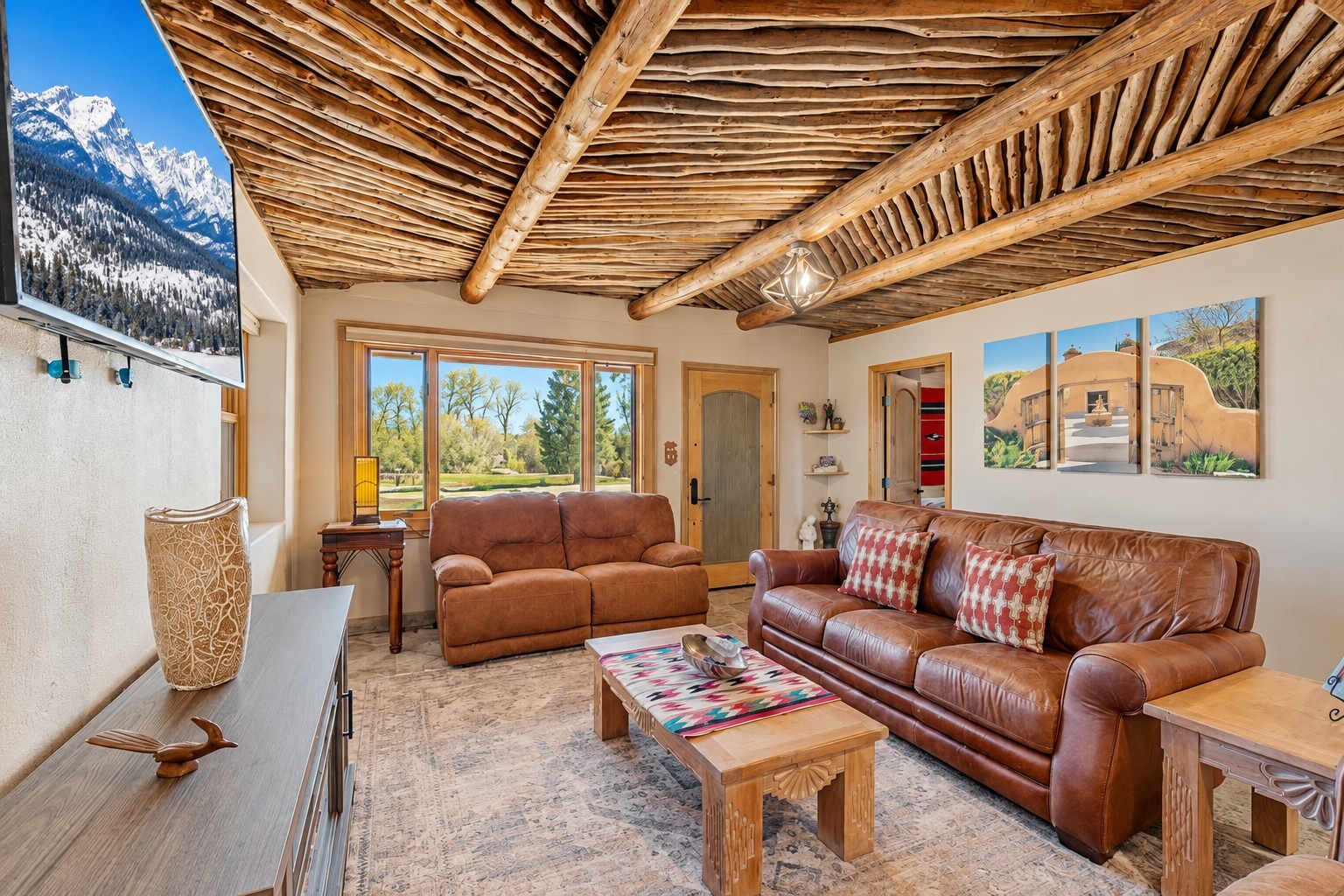 A cozy living room with a wooden ceiling, leather sofas, a coffee table, and a view of mountains through large windows.