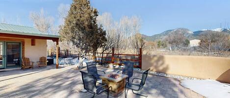 A patio with a fire pit table and chairs, a building with a green roof, and a view of mountains.