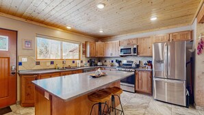 A kitchen with wooden cabinets, stainless steel appliances, a large island with stools, and a window providing natural light. - Casa de Rufina - Mountain Views, Firepit & Pets (Arroyo Seco)