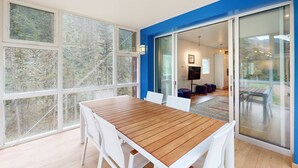 A modern interior with a wooden table and white chairs, large windows, and a glass door leading to an outdoor area. - Kachina Ridge Dreams - Walk to Lifts & Huge Views (Taos Ski Valley)
