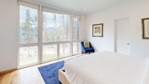 A bedroom with a large window, a bed, a blue chair, and framed pictures on the wall. - Kachina Ridge Dreams - Walk to Lifts & Huge Views (Taos Ski Valley)