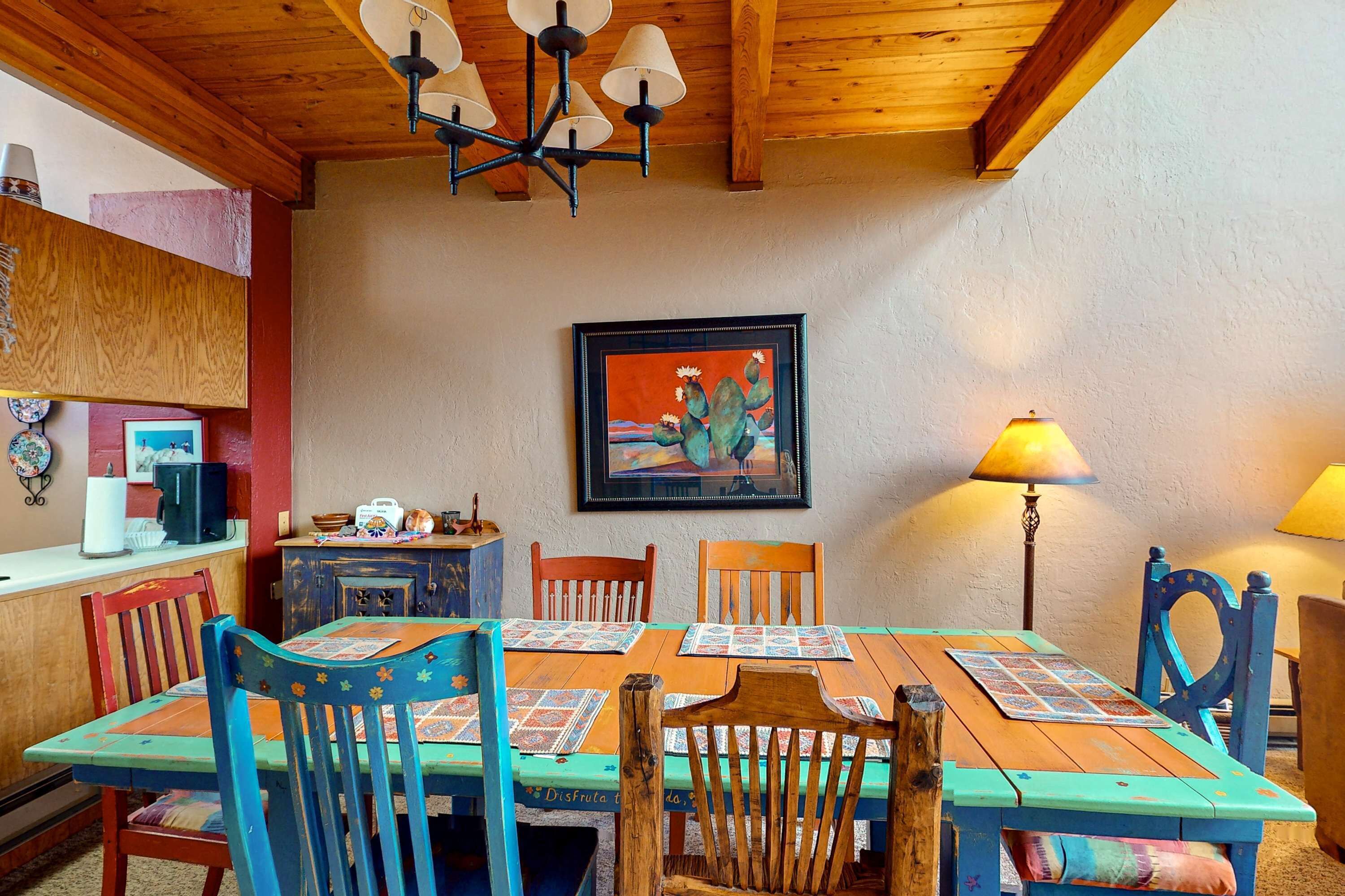 A dining area with wooden tables and chairs, a chandelier, a framed painting, and a lamp.