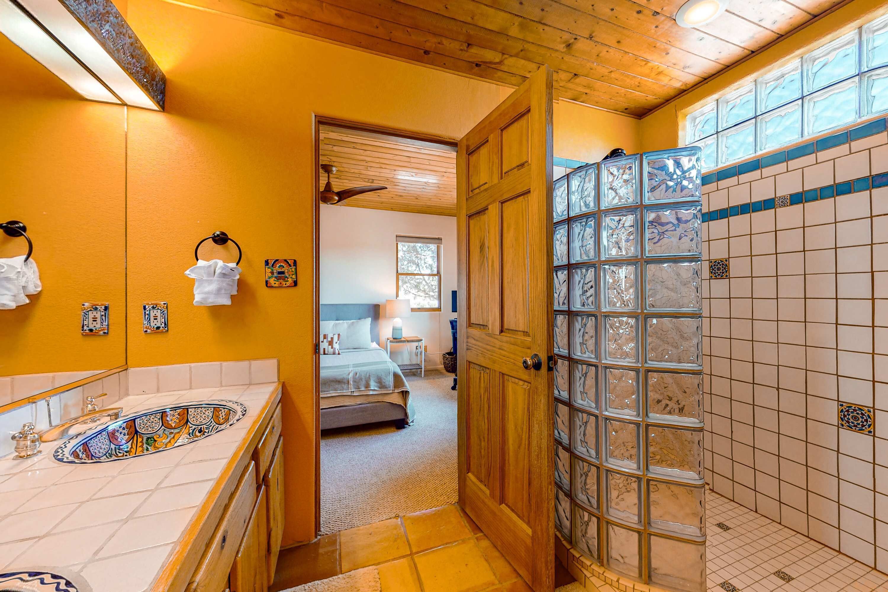 A bathroom with a wooden door, a tiled shower area, and a visible bedroom with a bed and window.
