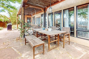 An outdoor patio with a wooden table and benches, surrounded by large windows and a pergola.