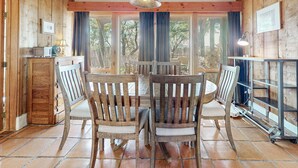 A wooden interior with a dining table and chairs, a hanging light fixture, and a window with curtains.
