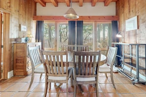 A wooden interior with a dining table and chairs, a hanging light fixture, and a window with curtains.