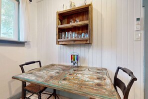 A dining table with chairs, a wooden shelf above with glassware, and a wall-mounted light switch.