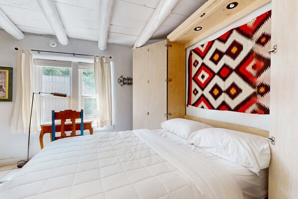 Une chambre à coucher comprenant un lit, une tête de lit en bois, une fenêtre avec des rideaux, une chaise et une œuvre d’art accrochée au mur.