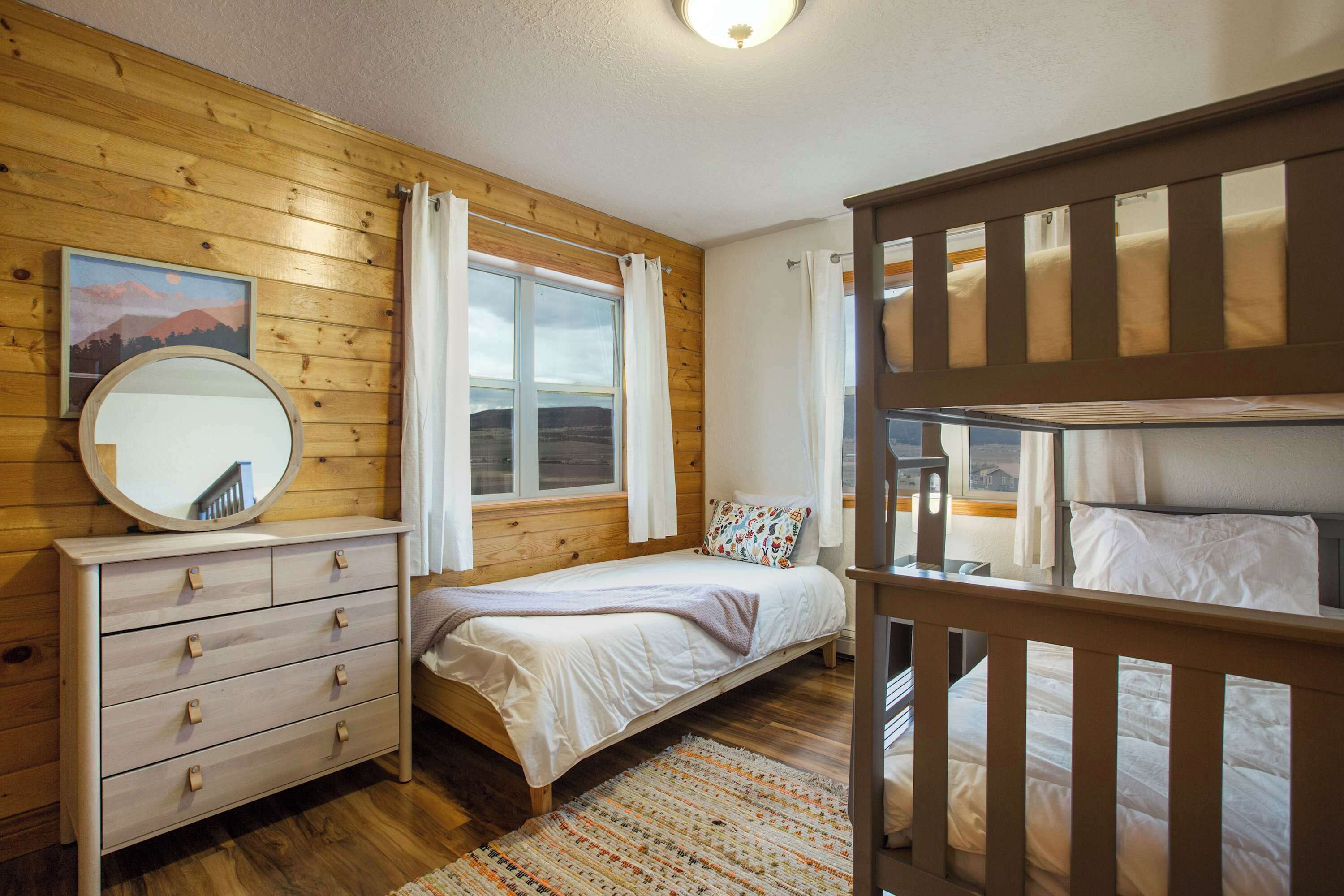 Une chambre avec des murs en bois, un lit, une commode, un miroir et une fenêtre avec des rideaux.