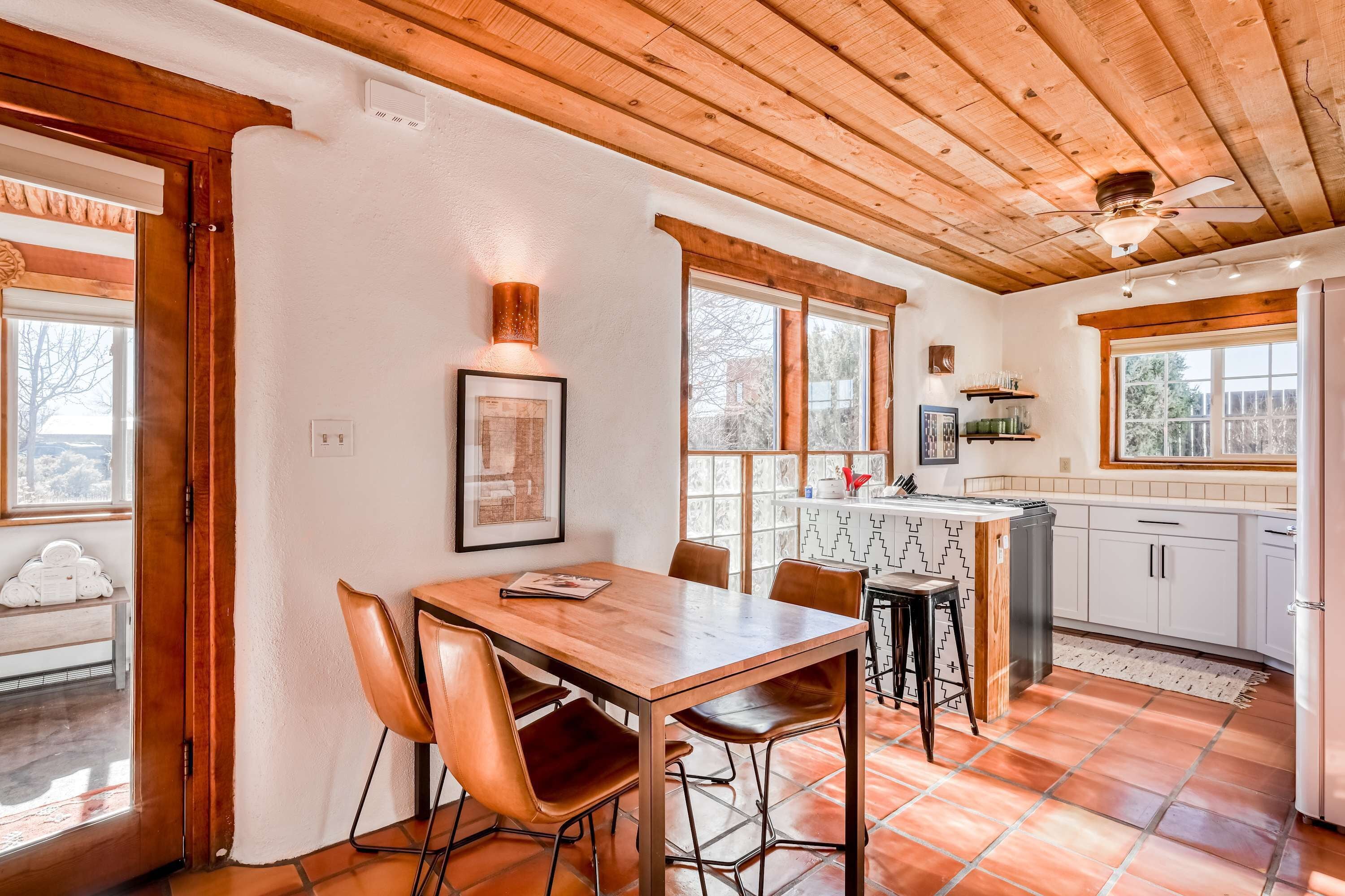 A dining area with a wooden table and chairs, a kitchen with white cabinets, and large windows.