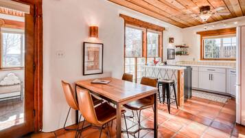 A dining area with a wooden table and chairs, a kitchen with white cabinets, and large windows.