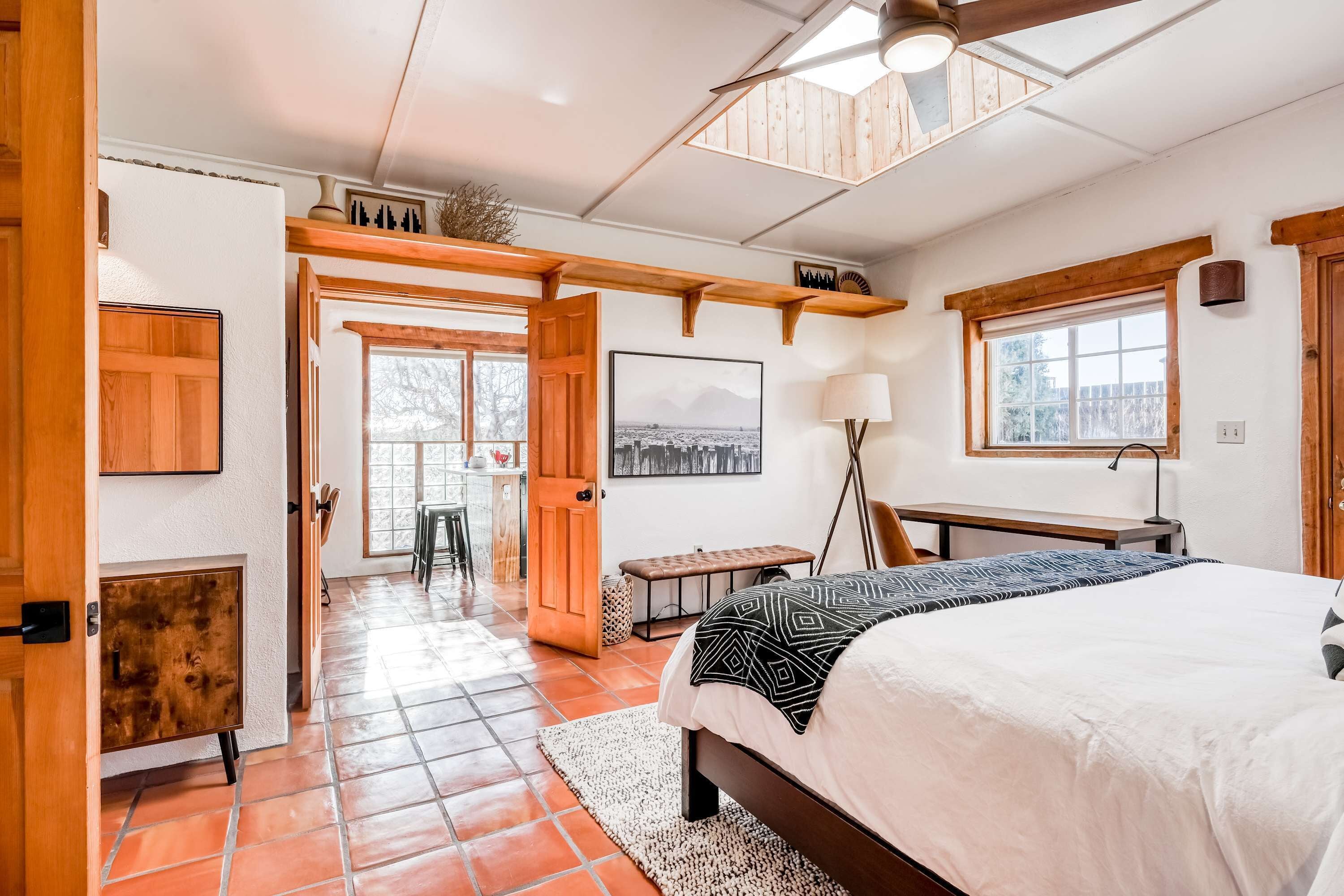 A bedroom with a bed, a desk, a chair, a lamp, a bench, and a door leading to another room.