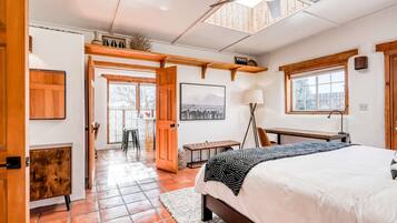 A bedroom with a bed, a desk, a chair, a lamp, a bench, and a door leading to another room.