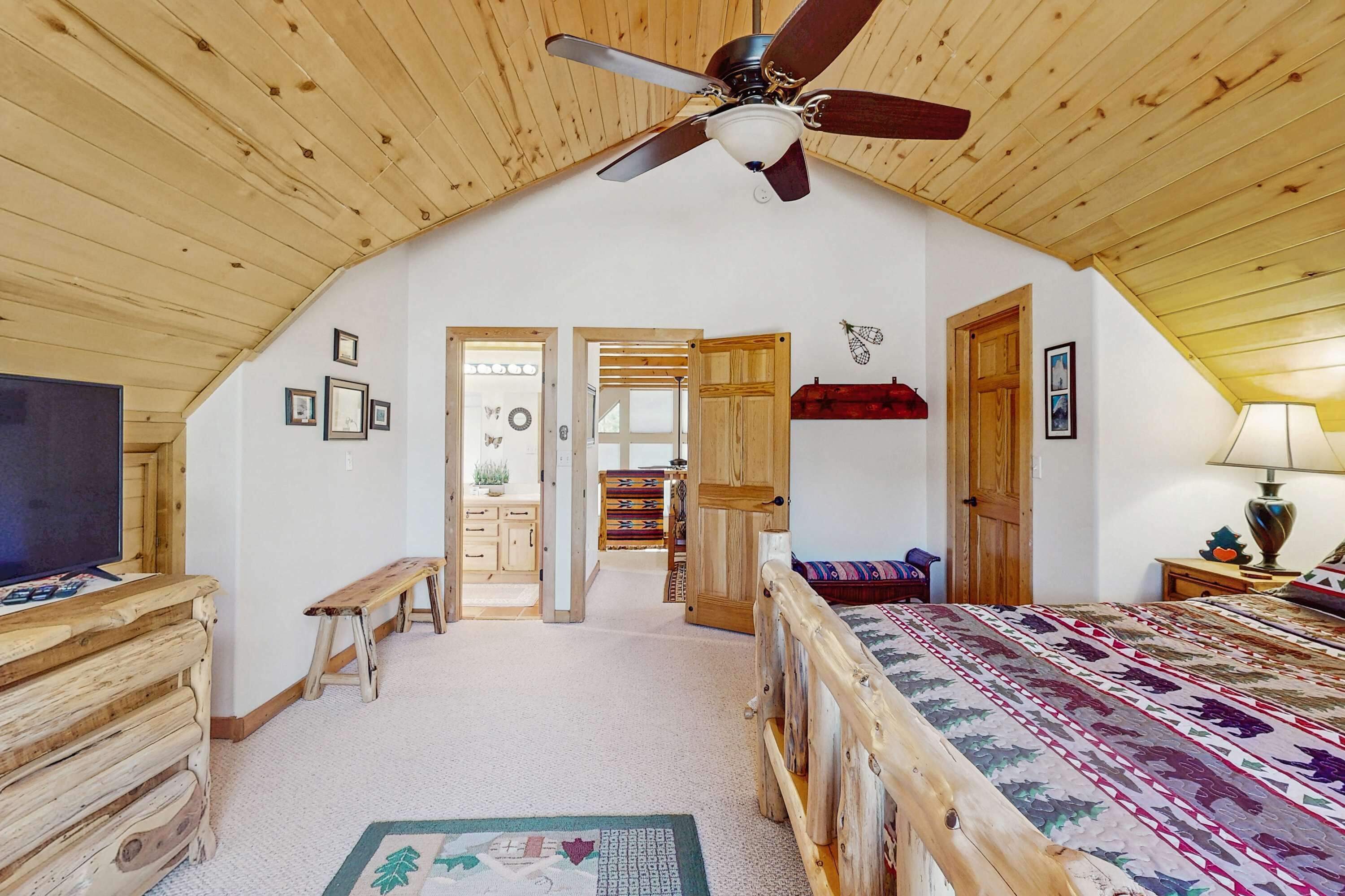 Une chambre avec un plafond en bois, un lit recouvert d’une couvre-lit à motifs, un ventilateur de plafond et un téléviseur posé sur une commode en bois.