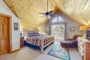 Ein Schlafzimmer mit Holzbalkendecke, einem Bett mit gemusterten Betttextilien, einem Schreibtisch mit Lampe und Blick ins Freie.
