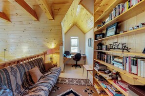 Ein gemütliches Blockhäuschen im Inneren mit Holzbalkendecke, einer Couch, einem Schreibtisch mit Computer und einem Bücherregal voller Bücher.