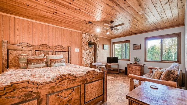 A wooden-themed bedroom with a bed, a TV, a stone wall, and a view of trees through large windows.
