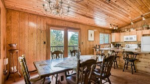 A wooden interior with a dining table, chairs, a chandelier, and a kitchen area.