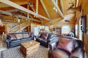 A log cabin interior with a wooden staircase, leather sofas, a wooden coffee table, and mounted animal heads.