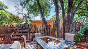 An outdoor seating area with a glass-top table and metal chairs, surrounded by trees and a wooden fence. - Casa Oeste - Walk to Plaza, Shaded Patio, Dog OK (Santa Fe)