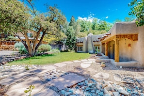 A courtyard with stone pathways, a tree, and a building with a unique architectural style. - Casa del Suenos - Steps to Plaza - Patio & Garden (Taos)