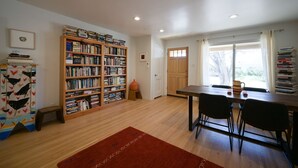 Une pièce avec une bibliothèque en bois, une table à manger avec des chaises, un petit tabouret et une fenêtre avec des rideaux.