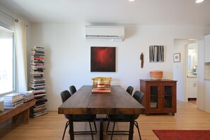 Ein Essbereich mit einem Holztisch, Stühlen, einem Bücherstapel, einer Klimaanlage und einem Bild an der Wand.
