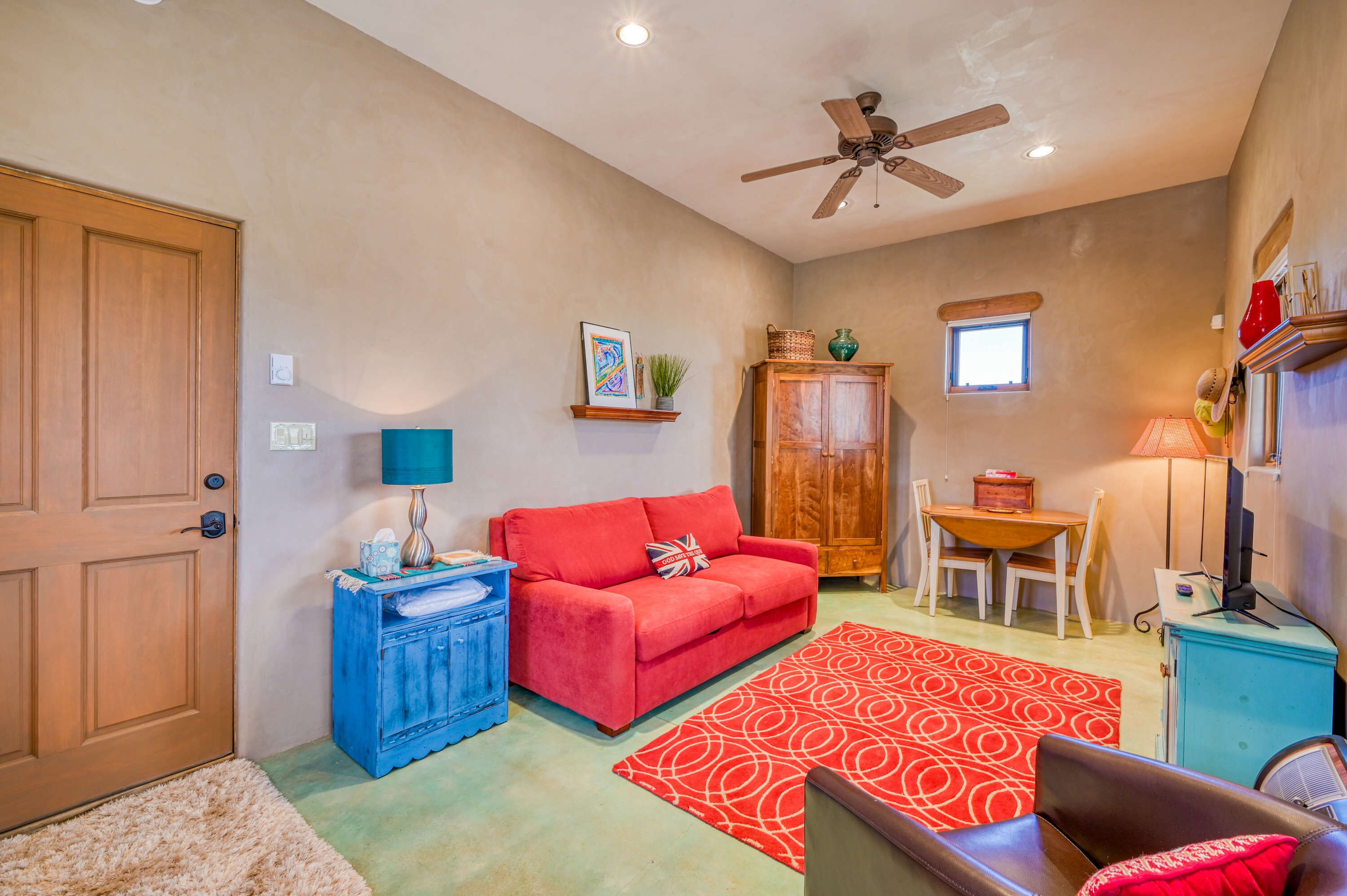 A room with a wooden door, a red sofa, a blue cabinet, a ceiling fan, a small table with chairs, and a window.