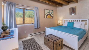 A bedroom with a bed, a bedside table, a window with curtains, a painting on the wall, and a wooden chest. - Medina Casita - Mountain Views, Firepit, Pets Okay (El Prado)