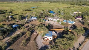 An aerial view of a rural property with multiple buildings, including a main house with a blue roof, surrounded by trees and open fields. - Medina Casita - Mountain Views, Firepit, Pets Okay (El Prado)