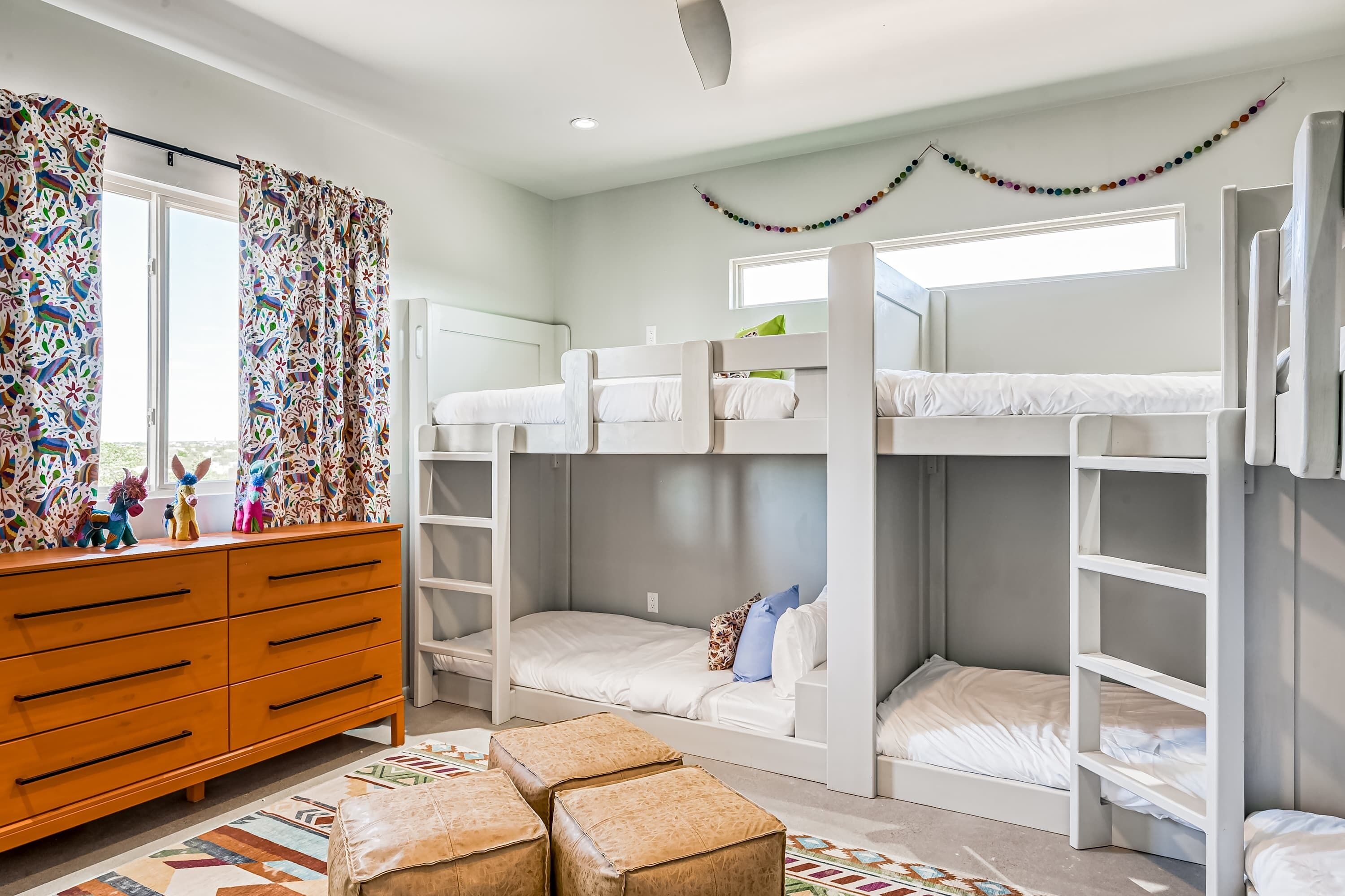 A bunk bed room with a dresser, a window with curtains, and a colorful wall decoration.