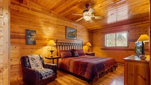 Ein Schlafzimmer in einer Blockhütte mit einem Bett, einem Sessel, einem Nachttisch, einer Lampe, einem Deckenventilator und einem Fenster.
