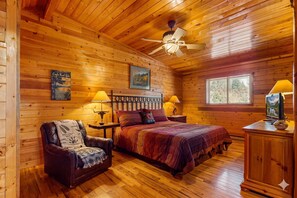 Un dormitorio tipo cabaña de madera con una cama, una silla, una mesita de noche, una lámpara, un ventilador de techo y una ventana.