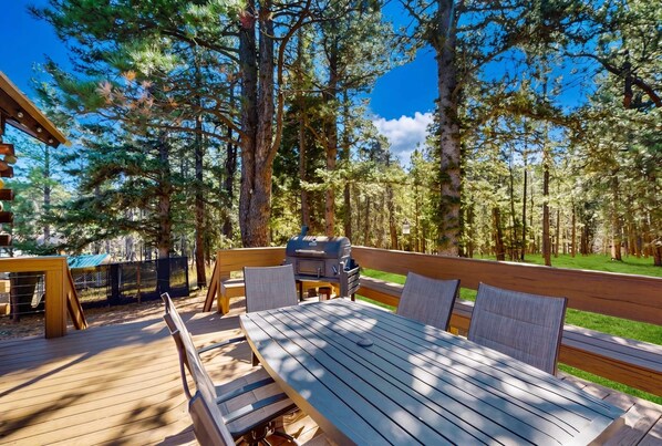 An outdoor deck with a table and chairs, surrounded by tall trees and a clear sky. - Angel Fire Big House - Mountain Views, Hike & Lake (Angel Fire)