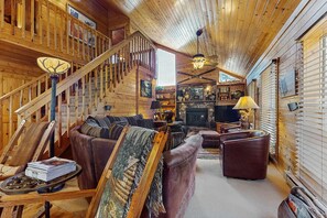 Interior de una cabaña de madera con una escalera, sofás de cuero, chimenea y televisor.