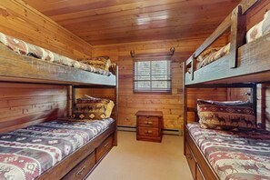 A cabin with wooden walls, two bunk beds with patterned bedding, a small wooden nightstand, and a window with blinds. - Angel Fire Big House - Mountain Views, Hike & Lake (Angel Fire)