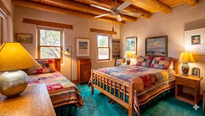 A bedroom with a wooden ceiling, a bed with a colorful quilt, two bedside tables with lamps, and framed pictures on the walls. - Adobe Retreat - Walk to Plaza, Fireplace, Patio (Taos)